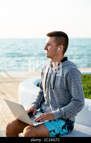 Lächelnd attraktiver Mann im Headset, ein Laptop verwenden, entfernt suchen, sitzt auf der Bank, am Kai. Im Freien. Stockfoto