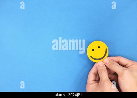 Glück und Positivität Konzept. Hand hält gelb lächelndes Gesicht in blauem Hintergrund mit Kopierraum. Stockfoto