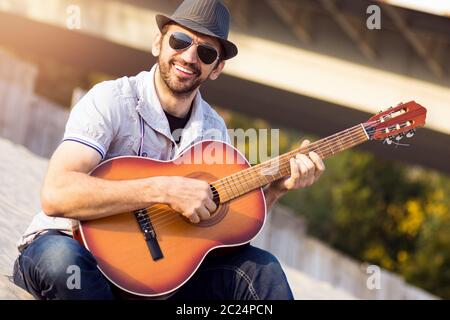 Der lächelnde modische Hipster Kerl in Hut und Sonnenbrille spielt Gitarre im Freien. Stockfoto