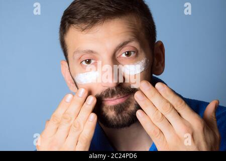 Gutaussehende Mann mittleren Alters ist mit Gesichtscreme auf seinem Gesicht auf blauem Hintergrund, Kosmetik und Hautpflege für Männer. Stockfoto