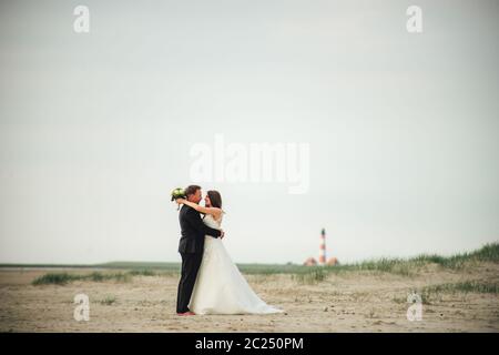 Brautpaar steht und umarmt am Sandstrand. Braut und Bräutigam schauen sich gegenseitig an und lächeln. Verschwommener Leuchtturm im Hintergrund. Hochzeitstag Stockfoto