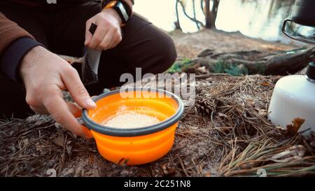 Das Thema des Tourismus ist Wandern und Reisen in der Natur. Die Hände Kaukasischer Mann gießt trocken getrockneten Fast-Food-Brei in einen Teller für CO Stockfoto