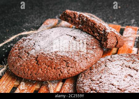 Zweieinhalb Schokoladenoatmeal-Kekse mit Puderzucker bestreut liegt auf einem Bambusplatte auf schwarzem Beton. Close up Stockfoto