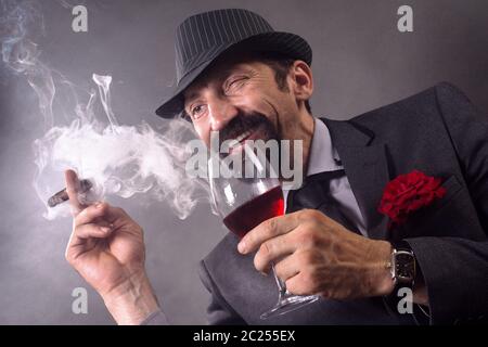 Der lächelnde zwinkende elegante Gentleman in Anzug und Hut raucht Zigarre und trinkt Wein auf grauem Hintergrund. Stockfoto