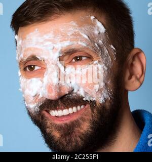 Der lächelnde Mann mittleren Alters verwendet Schaum für Hautpflege auf seinem Gesicht auf blauem Hintergrund. Stockfoto