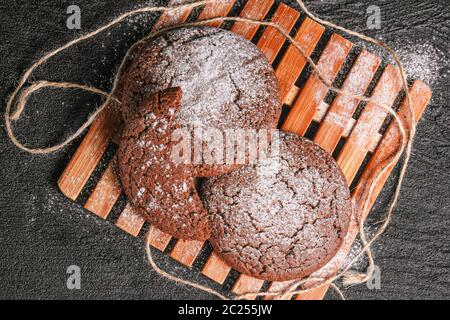 Zweieinhalb Schokoladenoatmeal-Kekse mit Puderzucker bestreut liegen auf einem Bambusplatte auf schwarzem Beton. Flach legen Draufsicht Stockfoto