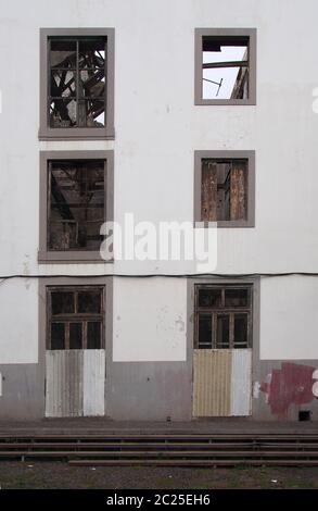 Die Fassade eines verlassenen großen weißen Industriegebäudes mit vertauften Türen und leeren Fenstern, die das zerstörte Dach abstellen Stockfoto