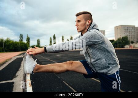 Close-up Profil einer jungen gutaussehenden Mann Übungen, Beine, das Hören von Musik über Kopfhörer, in Sportkleidung gekleidet, im Stadion Stockfoto