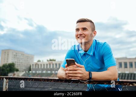 Lächelnd glückliche junge Menschen Musik hören, während sein Handy Holding, starrte in die Ferne, in lässig gekleidet, im Freien Stockfoto