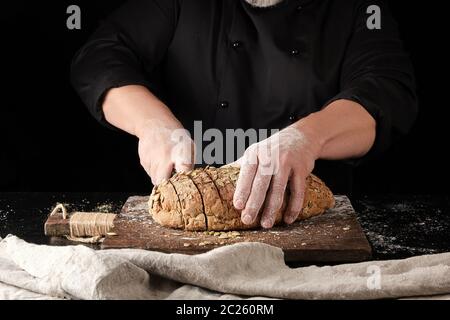 Baker in schwarzer Uniform Schnitte mit einem Messer in Scheiben von Roggen Brot mit Kürbiskernen auf braunem Holz- Board, schwarzer Hintergrund Stockfoto