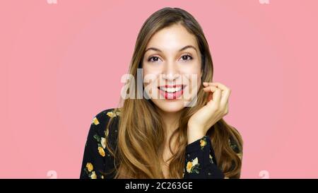 Lächelnd schöne junge Frau in schwarz geblümtem Kleid posiert mit Hand in der Nähe des Gesichts und Blick auf die Kamera über rosa Hintergrund Stockfoto