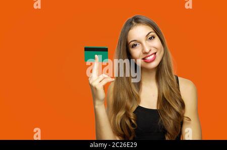 Junge Frau mit Kreditkarte in der Hand und lächelt in die Kamera, auf Orange Studio Hintergrund posiert Stockfoto