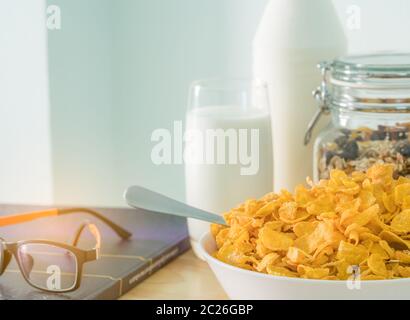 Schüssel Müsli mit Löffel und ein Glas Milch auf Holz Tisch in der Nähe von Diabetes Lehrbuch und Brillen. Calcium essen Frühstück. Cornflakes und Milch Stockfoto