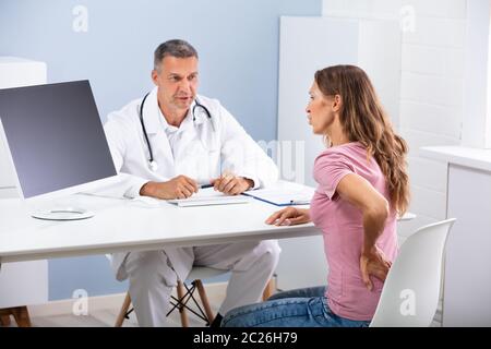 Männlicher Arzt im Gespräch mit weiblichen Patienten mit Rückenbeschwerden in der Klinik Stockfoto
