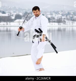 Mann in weißem Kimono übt Kampfkunst mit zwei Samurai-Schwertern barfuß auf Schnee im Winter. Stockfoto