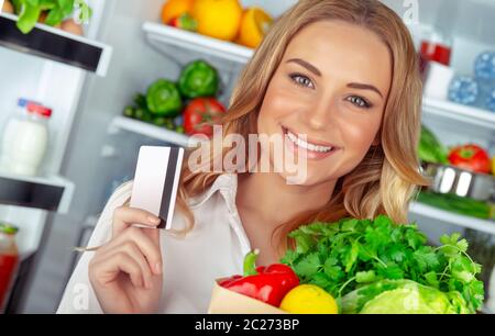 Käufer eines gesundes Essen, Porträt einer schönen Frau mit Kreditkarte und einer Papiertüte voll mit frischem Gemüse, in der Nähe von offenen Kühlschrank zu Hause stehen, org. Stockfoto