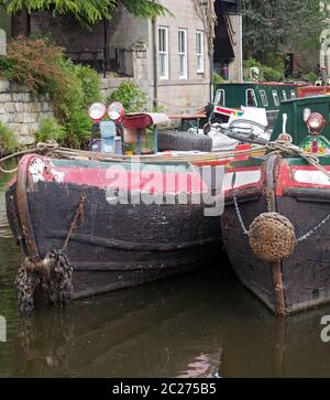 Die Bögen von zwei alten festgetäuten schmalen Boot auf dem rochdale Kanal in hebden Brücke von Grasbäumen und alten Gebäuden umgeben Stockfoto