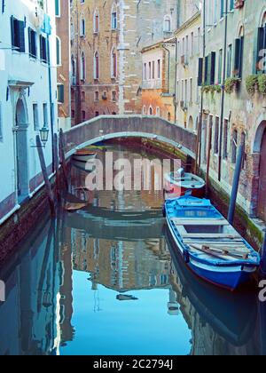 Ein ruhiger, enger Kanal, der von einer Brücke mit festgetäuten Booten und alten Gebäuden durchzogen wird, die sich im Wasser in Venedig spiegeln Stockfoto