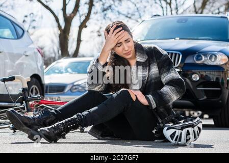 Volle Länge einer jungen Frau, die auf dem Asphalt mit schweren Kopfschmerzen und Schwindel nach einem Fahrradunfall auf einer belebten Straße der Stadt sitzt Stockfoto