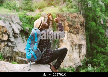 Afrikanischer Mann Reisende auf der Klippe sitzen und halten Kamera mit Rucksack Stockfoto