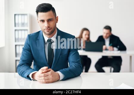 Porträt eines selbstbewussten, gutaussehenden Geschäftsmannes, der im Büro mit seinem Geschäftsteam auf dem Hintergrund sitzt Stockfoto