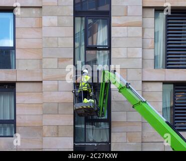 Ein nicht identifizierbarer Bauarbeiter in einer grünen Luftarbeitsbühne auf einer großen städtischen Wohnbaustelle Stockfoto