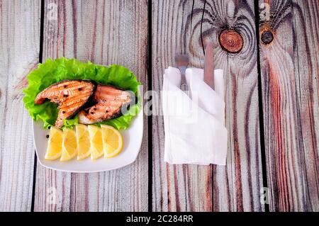 Zwei gegrillte rosafarbene Lachssteaks liegen auf einer weißen Keramikplatte, die mit Zitronenscheiben und einem Salatblatt neben einer weißen Serviette mit Besteck verziert ist, dem WH Stockfoto