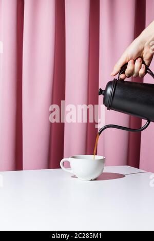 Frau Hand hält schwarzen Metalltopf und Gießen Kaffee oder Tee in weiße Tasse vor rosa Vorhänge, selektive Fokus Stockfoto