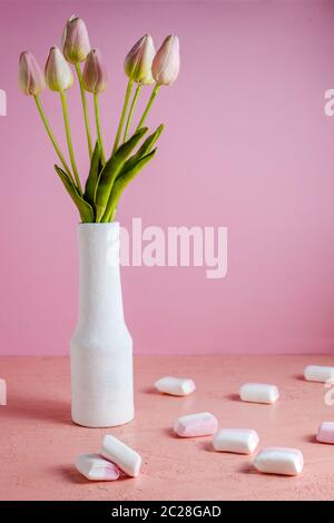 Copyspace mit bunten Mini Marshmallows auf einem Tisch neben rosa Tulpen in einer weißen Vase auf rosa Hintergrund. Stockfoto