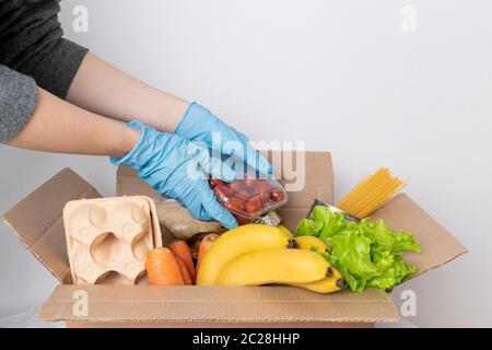 Box mit Futter zur Lieferung auf weißem Hintergrund. Eine weibliche Hand in einem medizinischen Handschuh steckt Kirschtomaten in eine Schachtel mit Produkten. Konzept der Spende, f Stockfoto