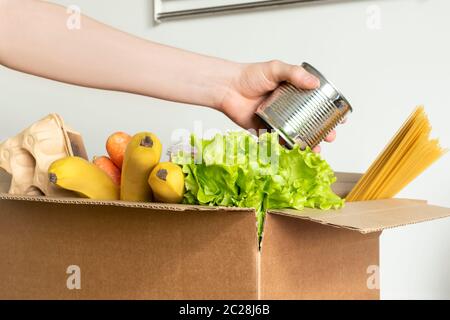 Box mit Futter zur Lieferung auf weißem Hintergrund. Die weibliche Hand in einem medizinischen Handschuh legt Dose in eine Schachtel mit Produkten. Konzept der Spende, Lieferung von Lebensmitteln Stockfoto