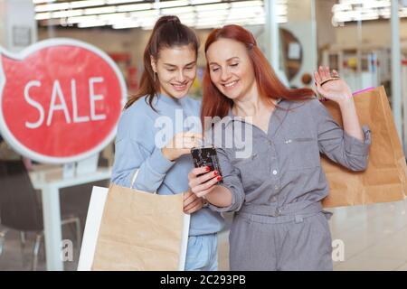 Zwei fröhliche Freundinnen lachen, benutzen Smartphone im Einkaufszentrum Stockfoto