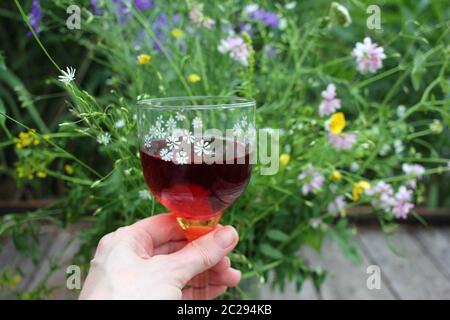 Ein Glas Rotwein mit Beeren in der Hand der Frau auf dem Hintergrund des verschwommenen Wildblumen-Bouquets. Mittsommernacht der Traum Erdbeer Cocktail Stockfoto