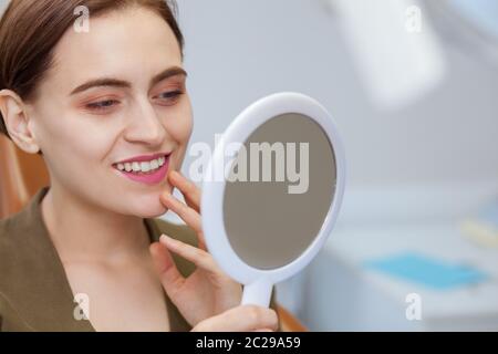 Nahaufnahme einer jungen schönen Frau lächelnd, Blick in den Spiegel in der Zahnklinik. Attraktive weibliche Patientin untersucht ihre perfekte weiß smil Stockfoto