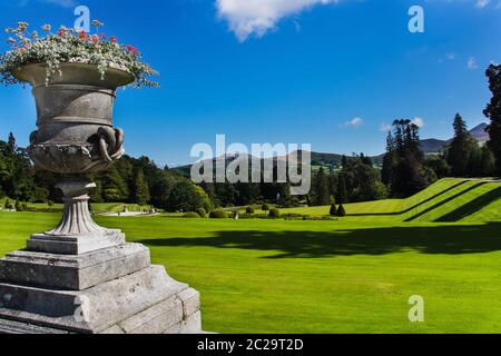 Eine reich verzierte Topf mit Blumen und breiten grünen Rasen in Powerscourt Gärten, Wald und Sugerloaf Berg im Hintergrund, Wicklow, Irland Stockfoto