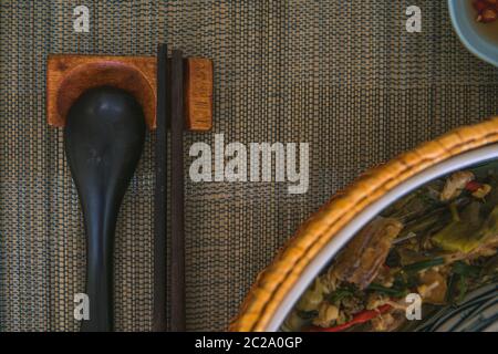 Nahaufnahme von Holzlöffel und Essstäbchen neben einem Rattan-Reis-Schüssel, innen ist ein traditionelles vietnamesisches Mittagessen, Reis, Suppe, salziges Essen. Stockfoto