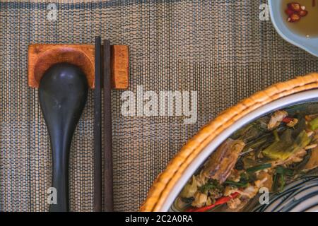 Nahaufnahme von Holzlöffel und Essstäbchen neben einem Rattan-Reis-Schüssel, innen ist ein traditionelles vietnamesisches Mittagessen, Reis, Suppe, salziges Essen. Stockfoto