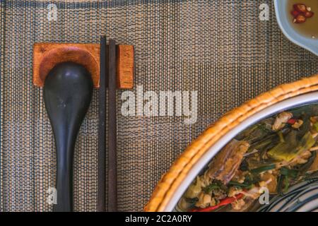 Nahaufnahme von Holzlöffel und Essstäbchen neben einem Rattan-Reis-Schüssel, innen ist ein traditionelles vietnamesisches Mittagessen, Reis, Suppe, salziges Essen. Stockfoto