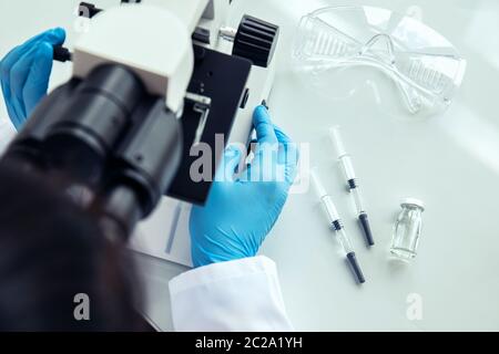 Wissenschaftlerin benutzt Mikroskop. Die Wissenschaftlerin in Schutzhandschuhen verwendet ein medizinisches Mikroskop mit Spritzen, Schutzbrille und Fläschchen daneben Stockfoto