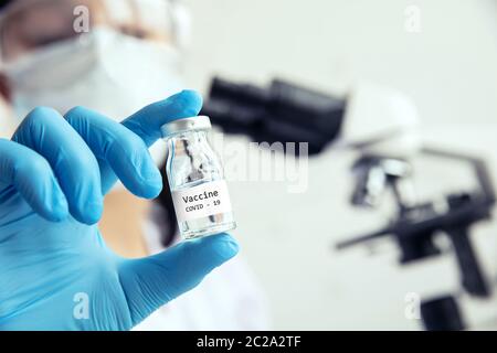 Hand in medizinischen Handschuhen hält Fläschchen mit Impfstoff. Die Medizinerin hält eine Fläschchen mit Impfstoff für Covid-19 in der Hand und ein Mikroskop im Hintergrund Stockfoto