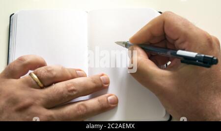 Männliche Hände halten einen Kugelschreiber, um auf die leeren Seiten eines offenen Notizbuchs zu schreiben.