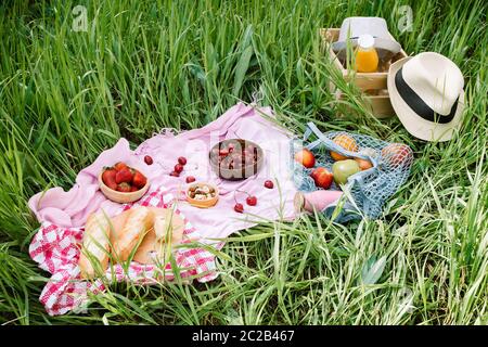 Zero Waste Sommer Picknick auf dem mit Kirschen in den hölzernen Kokosnussschalen, frischem Brot und Glasflasche Saft oder Smoothie auf rosa Decke, Flatlay Stockfoto