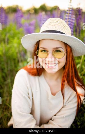 Junge rothaarige Mädchen mit langen Haaren in einem Weidenhut mit einem Band. Frau, die bei Sonnenuntergang auf einem Lupinenfeld sitzt und lächelt. Stockfoto