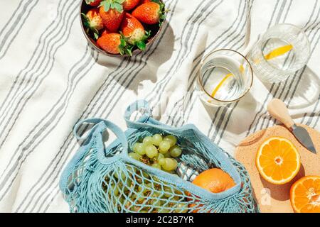 Feminine Sommer Picknick Flatlay, Früchte, String Öko-Bag und Zitronenwasser auf gestreiften Baumwolldecke Stockfoto