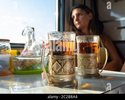 Auf dem Tisch im Abteil des Zuges sind Gläser mit Kaffee in Metall Becherhalter, im Hintergrund das Mädchen schaut aus dem Fenster Stockfoto