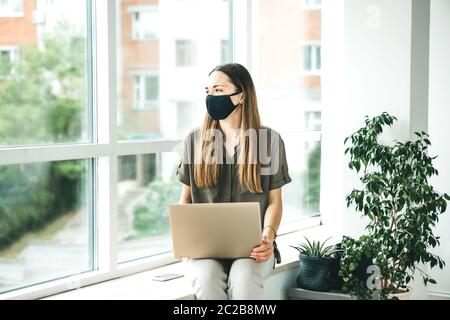 Traurige Arbeiterin oder Studentin, die mit einer Gesichtsmaske am Fenster mit einem Computer sitzt. Sie blieb zu Hause oder arbeitet am Fenster in einem offenen Raum. Stockfoto