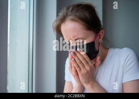 Frau trägt schwarze Gesichtsmaske, krank fühlen, leiden Niesen, Blick aus dem Fenster in Zimmer mit grauen Wand. Selbstisolierung, Prävention Stockfoto