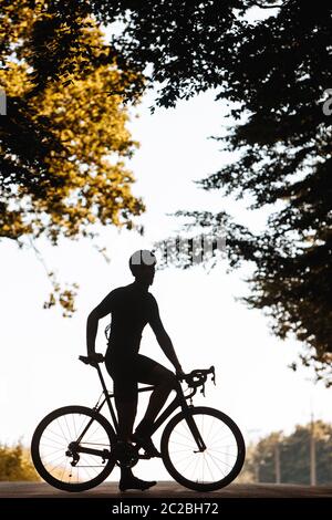 Starker Mann in Silhouette trägt Radbekleidung und Helm, posiert auf Kamera mit Fahrrad inmitten grüner Natur. Konzept der Selbstmotivation und Disziplin. Stockfoto