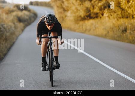 Bärtiger Radprofi im Sportoutfit und Schutzhelm aktiv auf asphaltierter Straße fahren. Aktiver Mann in gespiegelten Brillen Training unter grünen n Stockfoto