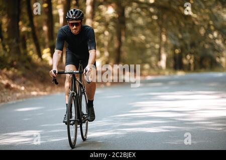 Reifer Mann in Sportbekleidung, Schutzhelm und Sonnenbrille Reiten schwarzen Profi-Fahrrad mit Unschärfe Hintergrund der Natur. Männliche Radfahrer genießen su Stockfoto